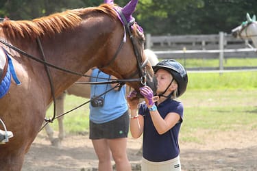 A child wearing a helmet and gloves kisses a horse's nose outdoors, showcasing the joys of choosing a camp that fosters connections with animals. Another person and a partially visible fence are in the background. riding camp horse