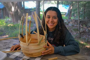 A person sits at a wooden table outdoors, smiling at the camera while engaging in art activities, specifically weaving a basket with wooden strips. Clothespins are attached to the basket, securing some of the strips in place. arts and crafts